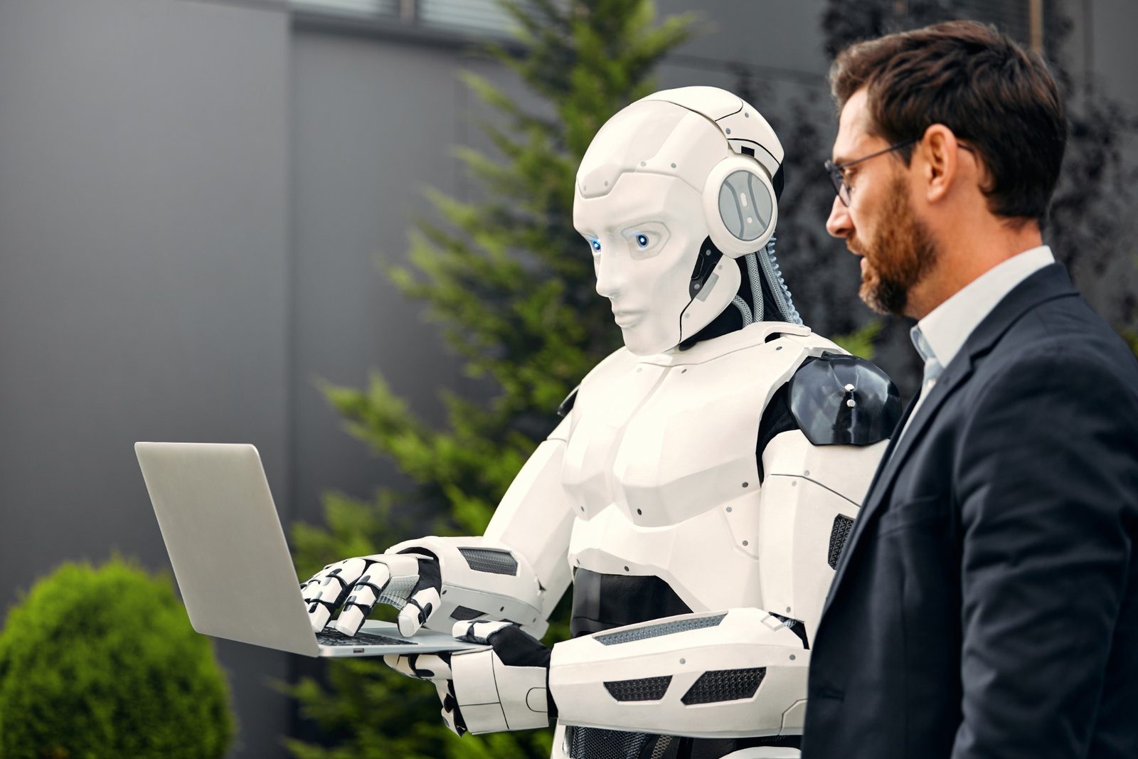 Humanoid robot typing on a laptop next to a businessman in a suit